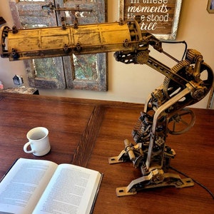 Puede incluir: Una l&aacute;mpara de estilo steampunk con una pantalla de metal amarilla y engranajes y palancas intrincados. La l&aacute;mpara est&aacute; sobre una mesa de madera con un libro y una taza de caf&eacute;.
