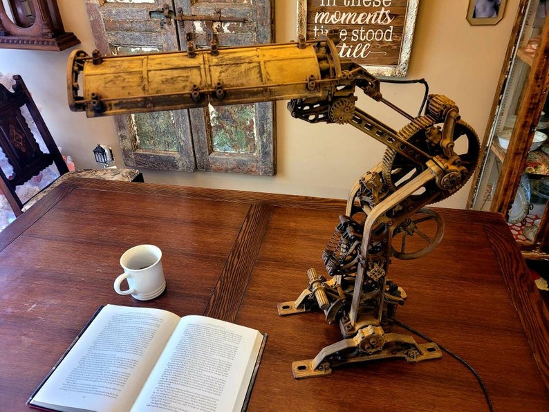 Puede incluir: Una l&aacute;mpara de estilo steampunk con una pantalla de metal amarilla y engranajes y palancas intrincados. La l&aacute;mpara est&aacute; sobre una mesa de madera con un libro y una taza de caf&eacute;.