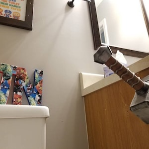 May include: A bathroom with a white toilet and a wooden cabinet. The cabinet has a mirror above it. A wooden letter "J" and "N" are on the toilet tank. A gray and brown Mjolnir hammer is mounted on the cabinet. The hammer has the word "Thor" written on it.