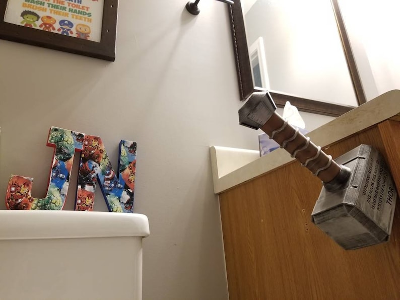 May include: A bathroom with a white toilet and a wooden cabinet. The cabinet has a mirror above it. A wooden letter "J" and "N" are on the toilet tank. A gray and brown Mjolnir hammer is mounted on the cabinet. The hammer has the word "Thor" written on it.