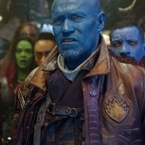 May include: A blue-skinned character with a shaved head and a red stripe on his forehead. He is wearing a brown leather jacket with a patch on the left side. He is surrounded by other characters.