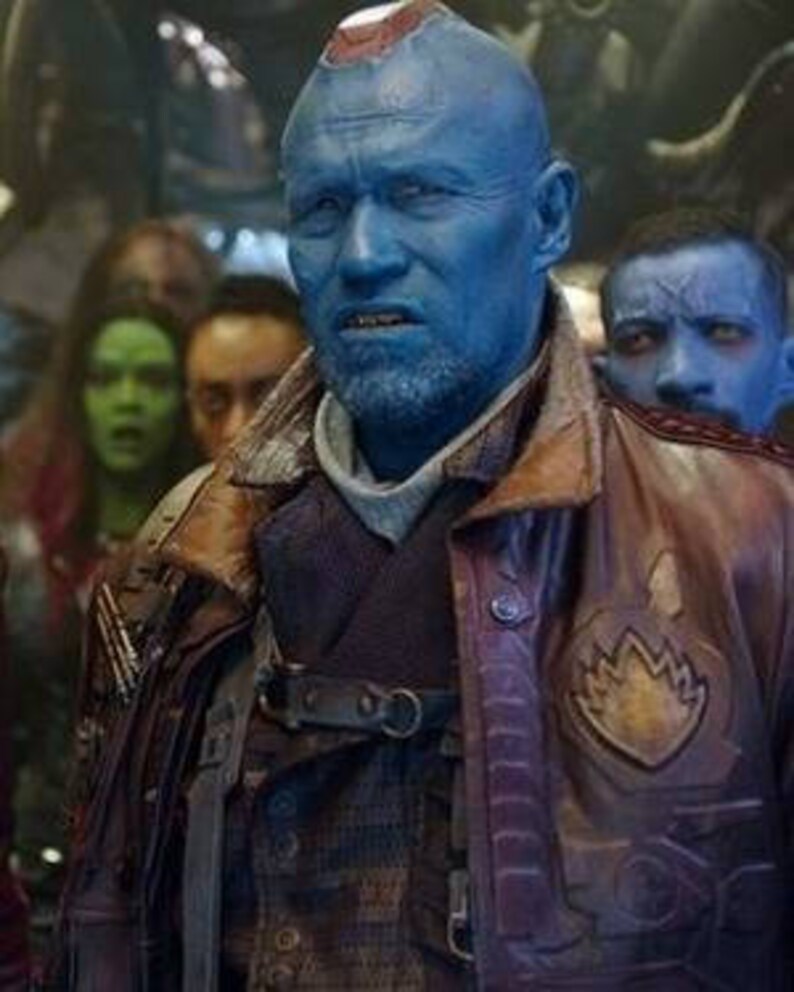 May include: A blue-skinned character with a shaved head and a red stripe on his forehead. He is wearing a brown leather jacket with a patch on the left side. He is surrounded by other characters.