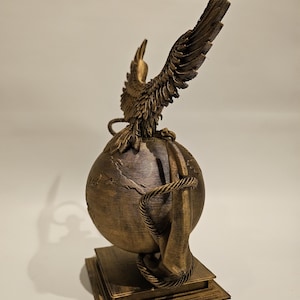 May include: A bronze-colored sculpture featuring a globe with a detailed map, encircled by a rope. An eagle with outstretched wings sits atop the globe. The sculpture is mounted on a square base.