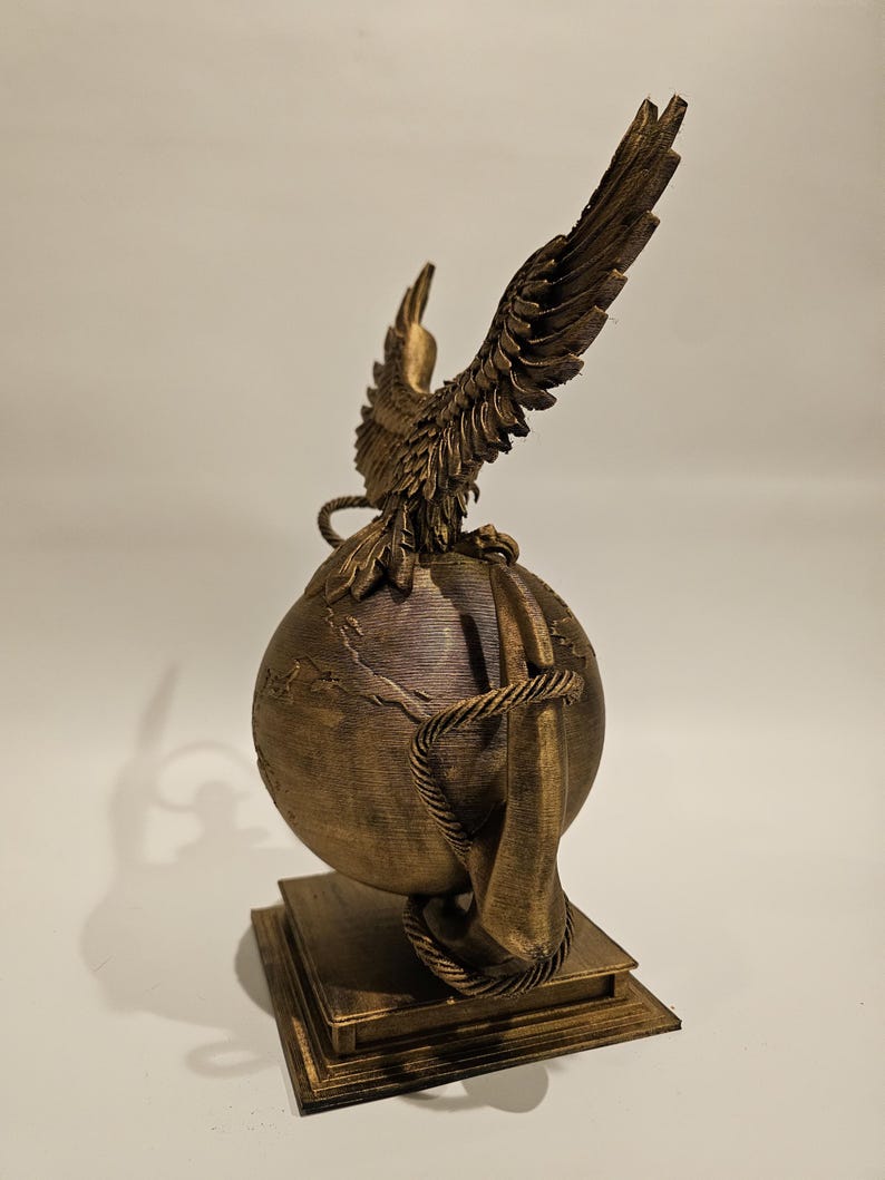 May include: A bronze-colored sculpture featuring a globe with a detailed map, encircled by a rope. An eagle with outstretched wings sits atop the globe. The sculpture is mounted on a square base.