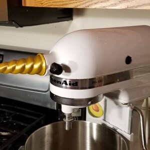 May include: A white KitchenAid stand mixer with a gold unicorn horn attached to the side. The mixer has a silver bowl and a silver control panel with the KitchenAid logo and the text "Pro 500" on it.