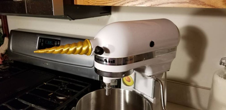 May include: A white KitchenAid stand mixer with a gold unicorn horn attached to the side. The mixer has a silver bowl and a silver control panel with the KitchenAid logo and the text "Pro 500" on it.