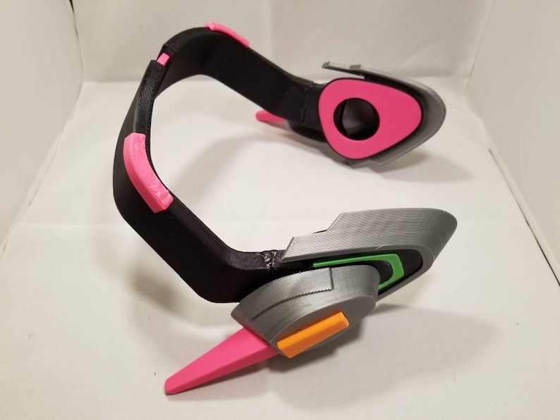 Puede incluir: Una diadema negra impresa en 3D con detalles en rosa y verde. La diadema tiene un auricular gris y verde con un detalle naranja.
