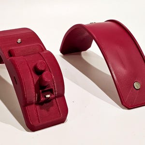 Op de afbeelding: Twee 3D-geprinte rode kunststof onderdelen voor een project. Een onderdeel is een gebogen stuk met een vlak oppervlak. Het andere onderdeel is een gebogen stuk met een verhoogd gedeelte met twee ronde knoppen en een klein rechthoekig gleuf.