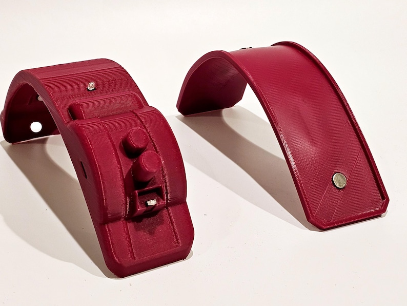 Op de afbeelding: Twee 3D-geprinte rode kunststof onderdelen voor een project. Een onderdeel is een gebogen stuk met een vlak oppervlak. Het andere onderdeel is een gebogen stuk met een verhoogd gedeelte met twee ronde knoppen en een klein rechthoekig gleuf.
