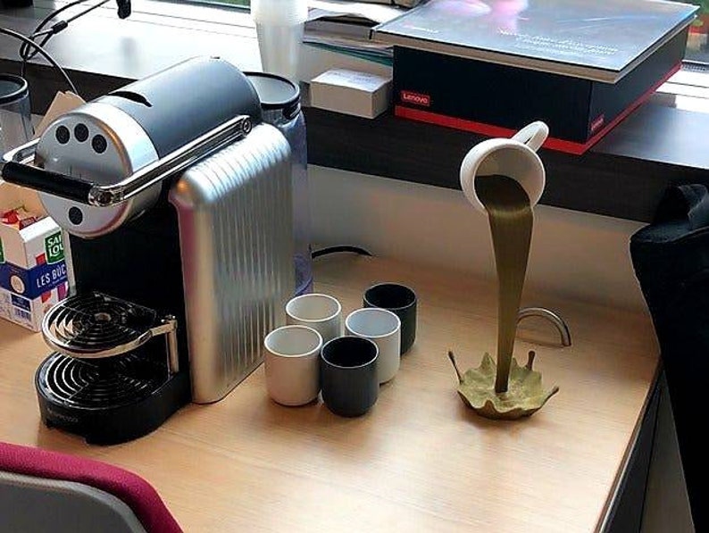 May include: A black and silver espresso machine with a drip tray and a cup holder. Four white ceramic mugs are on the counter, and a white ceramic cup is pouring a brown liquid into a gold-colored splash.