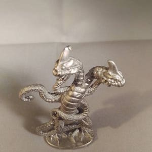 May include: A silver three-headed dragon miniature figurine. The dragon has a detailed, scaly body and three heads with open mouths.