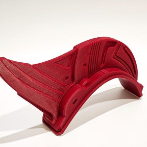 Op de afbeelding: Een rood 3D-geprint plastic object met een gebogen vorm en geometrische ontwerpen. Het object heeft een scherpe punt aan het ene uiteinde en een afgeronde rand aan het andere uiteinde.