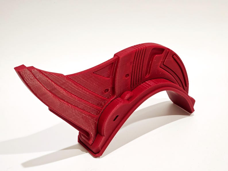Op de afbeelding: Een rood 3D-geprint plastic object met een gebogen vorm en geometrische ontwerpen. Het object heeft een scherpe punt aan het ene uiteinde en een afgeronde rand aan het andere uiteinde.