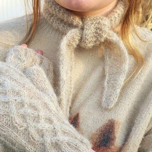 May include: A close-up of a person wearing a light brown knitted sweater with a matching knitted scarf and gloves. The sweater has a loose fit and a cable knit pattern. The scarf is tied in a loose knot around the neck. The gloves are fingerless and have a similar cable knit pattern to the sweater.