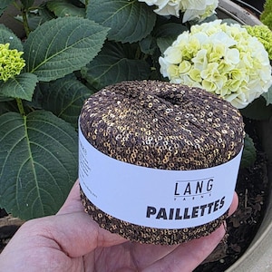 May include: A brown and gold sequined yarn ball with a white label that reads "LANG YARNS PAILLETTES". The yarn is held in a hand, with green leaves and white and yellow flowers in the background.