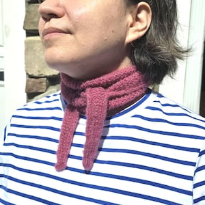May include: A close-up of a person wearing a pink knitted scarf. The scarf is wrapped around the neck and has two long, dangling ends. The person is wearing a white and blue striped shirt.