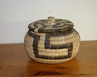 May include: A round, woven basket with a lid. The basket is made of natural materials and has a brown and white geometric pattern.