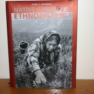 May include: A book titled "Native American Ethnobotany" by Daniel E. Moerman. The cover features a black and white photograph of a woman in a headscarf, kneeling in a field of plants.
