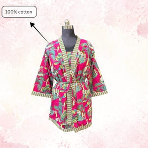 May include: A vibrant pink cotton robe with a tropical print featuring green leaves and animal figures. The robe has a striped green and white trim, a matching belt, and the text "100% cotton".