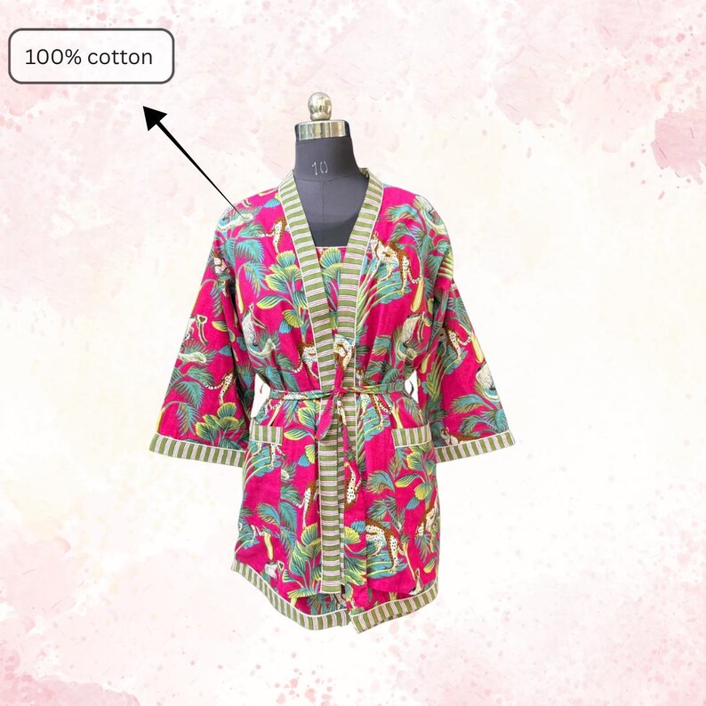 May include: A vibrant pink cotton robe with a tropical print featuring green leaves and animal figures. The robe has a striped green and white trim, a matching belt, and the text "100% cotton".