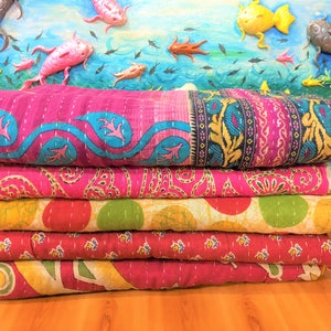 May include: A stack of colorful, patterned quilts. The top quilt is a vibrant pink with blue and white accents. The other quilts feature red, yellow, and green patterns. The background has a painted ocean scene with fish.