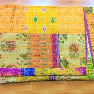 Peut inclure: Une courtepointe pliée, composée de différents tissus aux couleurs vives. Elle présente des sections jaunes, oranges et à motifs, cousues avec des points blancs. Des motifs floraux et géométriques créent une esthétique artisanale et chaleureuse.