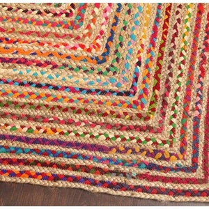 May include: Braided jute rug with a vibrant, colorful design. The rug features a natural beige base with interwoven bands of red, orange, purple, blue, and green. The edges are neatly finished, creating a geometric pattern.