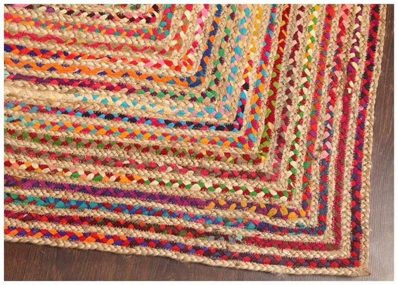 May include: Braided jute rug with a vibrant, colorful design. The rug features a natural beige base with interwoven bands of red, orange, purple, blue, and green. The edges are neatly finished, creating a geometric pattern.