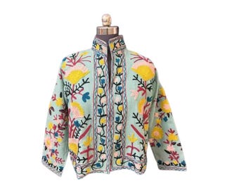 May include: A light blue jacket with colorful floral embroidery. The jacket features yellow, pink, blue, and red flower designs. The jacket has a high collar and is displayed on a mannequin.