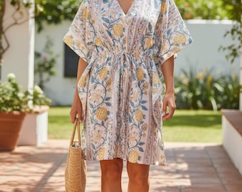 May include: A V-neck dress with a floral pattern in yellow and blue on a white background. The dress has short sleeves and a drawstring waist. A straw tote bag and white shoes complete the look.