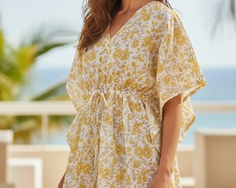 May include: A yellow floral print dress with a V-neck and drawstring waist. The dress has short, wide sleeves and falls to mid-thigh. The background shows a beach scene with palm trees and a blue ocean.