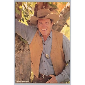 May include: A vintage postcard featuring a smiling man in a cowboy hat and vest. He wears a light blue shirt, a tan vest, and a brown belt. A badge is pinned on the vest. The text "Marshal Matt Dillon" is at the bottom.