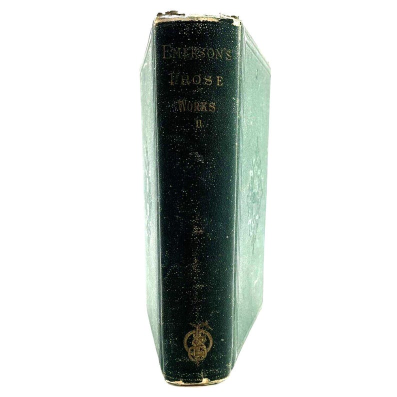 May include: An antique book with a dark green cover and gold lettering. The title "Emerson's Prose Works" is visible. The book's spine shows signs of wear, indicating age. A decorative emblem is at the bottom of the spine.