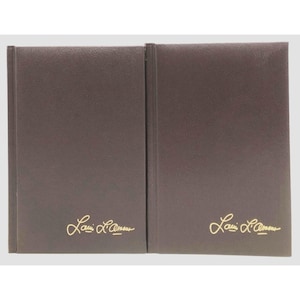May include: Two brown leather-bound books with gold lettering that reads "Lavi L'Amour". The books are side-by-side, with a vertical spine on the left side of each book. The books are on a light gray background.
