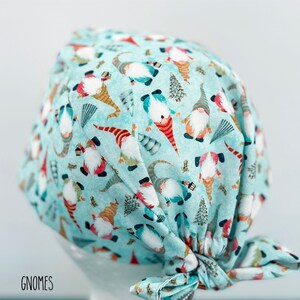 May include: A light blue surgical scrub cap with a repeating pattern of gnomes, trees, and other festive elements. The gnomes have red, orange, and blue hats and clothing. The word "GNOMES" is printed in black at the bottom left.