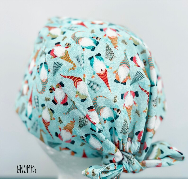 May include: A light blue surgical scrub cap with a repeating pattern of gnomes, trees, and other festive elements. The gnomes have red, orange, and blue hats and clothing. The word "GNOMES" is printed in black at the bottom left.