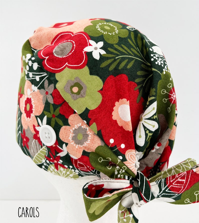 May include: A floral patterned scrub cap with a bow. The cap features a dark green background with red, pink, and white flowers. A white button is on the side. The word "CAROLS" is printed at the bottom.