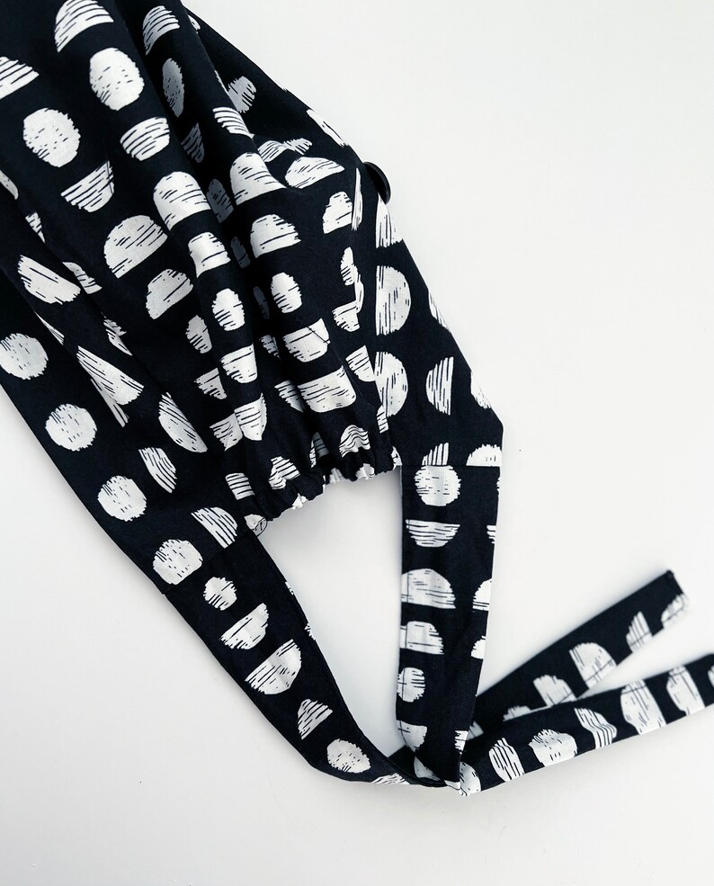 May include: Black and white patterned fabric scrub cap with two long ties. The fabric has a geometric pattern of circles and semi-circles.