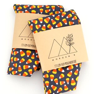 May include: Two folded fabric items with a dark gray background covered in a pattern of candy corn. Each item is wrapped with a brown paper band featuring the text "MORGAN K" and "etsy.com/shop/MORGANBOUTIQUE".