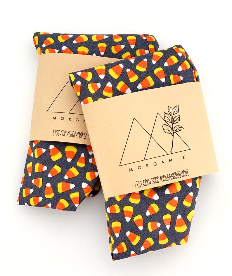 May include: Two folded fabric items with a dark gray background covered in a pattern of candy corn. Each item is wrapped with a brown paper band featuring the text "MORGAN K" and "etsy.com/shop/MORGANBOUTIQUE".