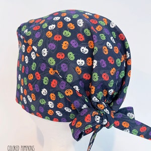 May include: A navy blue surgical scrub cap featuring a colorful pumpkin pattern in orange, purple, green, and white. The cap is tied at the back with a matching fabric tie. The text "COLORED PUMPKINS" is printed at the bottom.