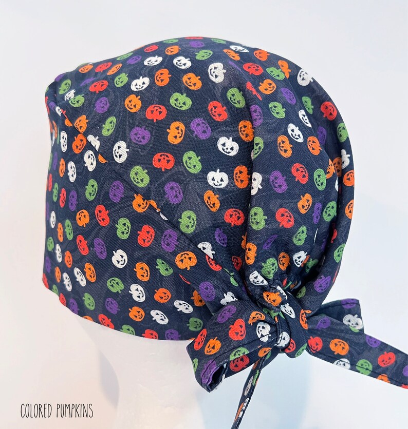 May include: A navy blue surgical scrub cap featuring a colorful pumpkin pattern in orange, purple, green, and white. The cap is tied at the back with a matching fabric tie. The text "COLORED PUMPKINS" is printed at the bottom.