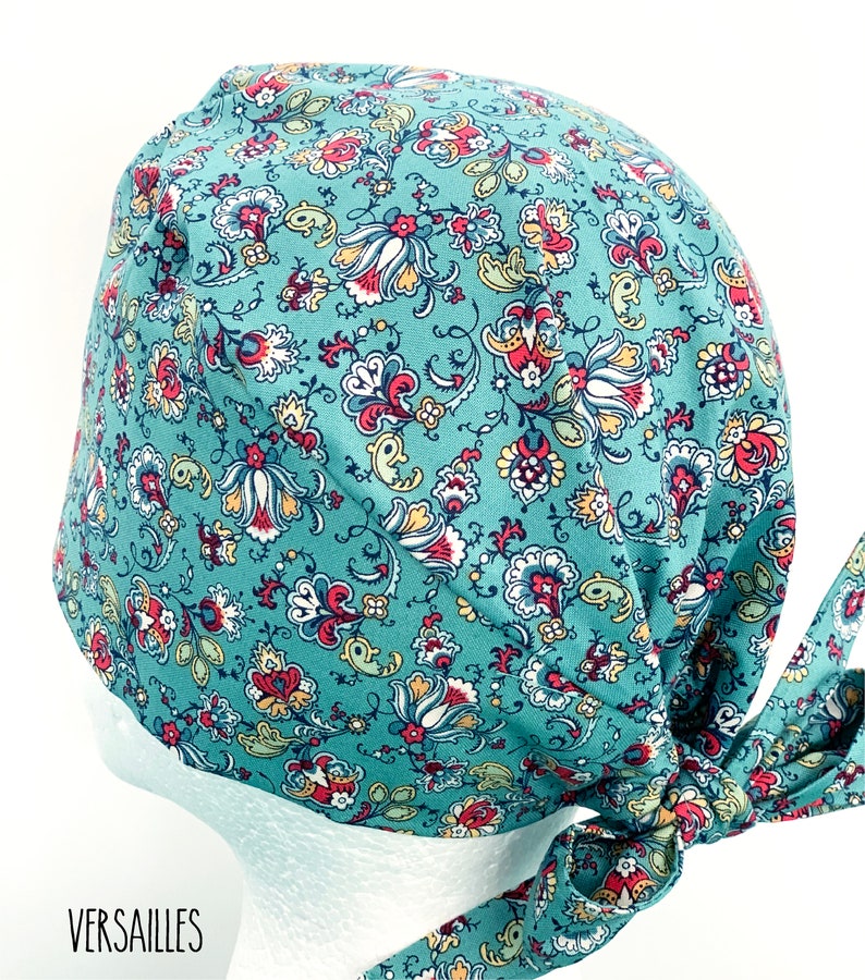 K&ouml;nnte beinhalten: Eine tealblaue OP-M&uuml;tze mit einem floralen Muster in Rot-, Gelb- und Wei&szlig;t&ouml;nen. Die M&uuml;tze hat einen Bindfaden an der R&uuml;ckseite.