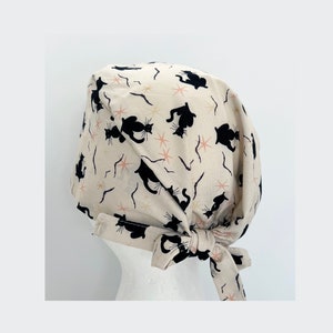 May include: A beige surgical scrub cap featuring a repeating pattern of black cat silhouettes, pink starbursts, and curved lines. The cap is tied at the back with a matching fabric tie. The cap is designed to be worn in a medical setting.