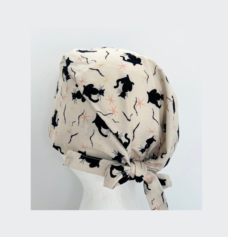 May include: A beige surgical scrub cap featuring a repeating pattern of black cat silhouettes, pink starbursts, and curved lines. The cap is tied at the back with a matching fabric tie. The cap is designed to be worn in a medical setting.