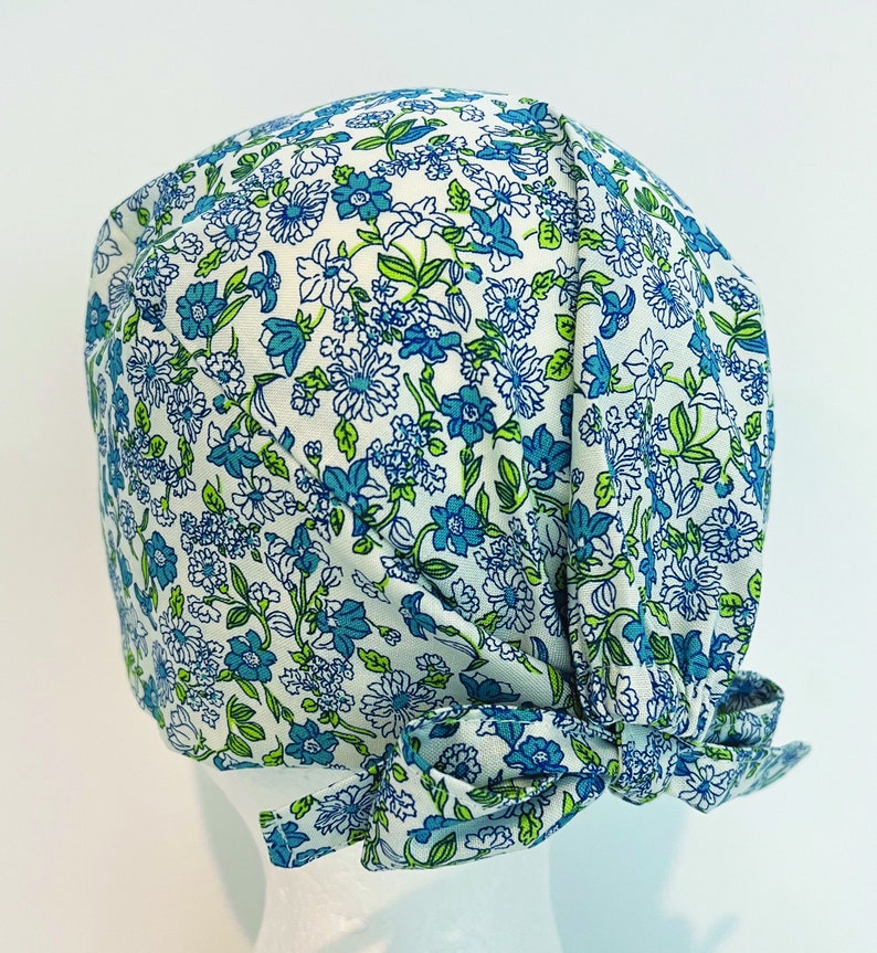 K&ouml;nnte beinhalten: Eine wei&szlig;e OP-M&uuml;tze mit einem blauen und gr&uuml;nen Blumenmuster. Die M&uuml;tze hat einen Knoten am R&uuml;cken.