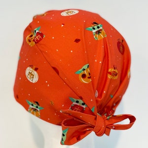 May include: Orange surgical scrub cap featuring a repeating pattern of Baby Yoda characters in Halloween costumes and pumpkins. The cap has a tie closure and the text "Mandalorian Halloween" is printed on the front.
