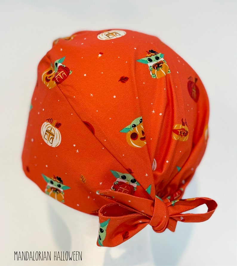 May include: Orange surgical scrub cap featuring a repeating pattern of Baby Yoda characters in Halloween costumes and pumpkins. The cap has a tie closure and the text "Mandalorian Halloween" is printed on the front.