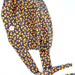 May include: A dark gray surgical scrub cap with a repeating pattern of candy corn in orange, yellow, and white. The cap has a white button and long ties for securing. The fabric is gathered at the back.