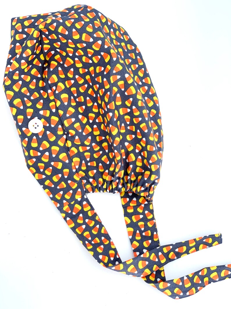 May include: A dark gray surgical scrub cap with a repeating pattern of candy corn in orange, yellow, and white. The cap has a white button and long ties for securing. The fabric is gathered at the back.
